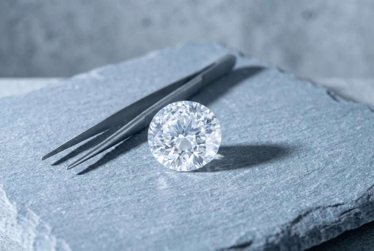 A single large loose round diamond alone on a gray stone plinth beside a pair of tweezers