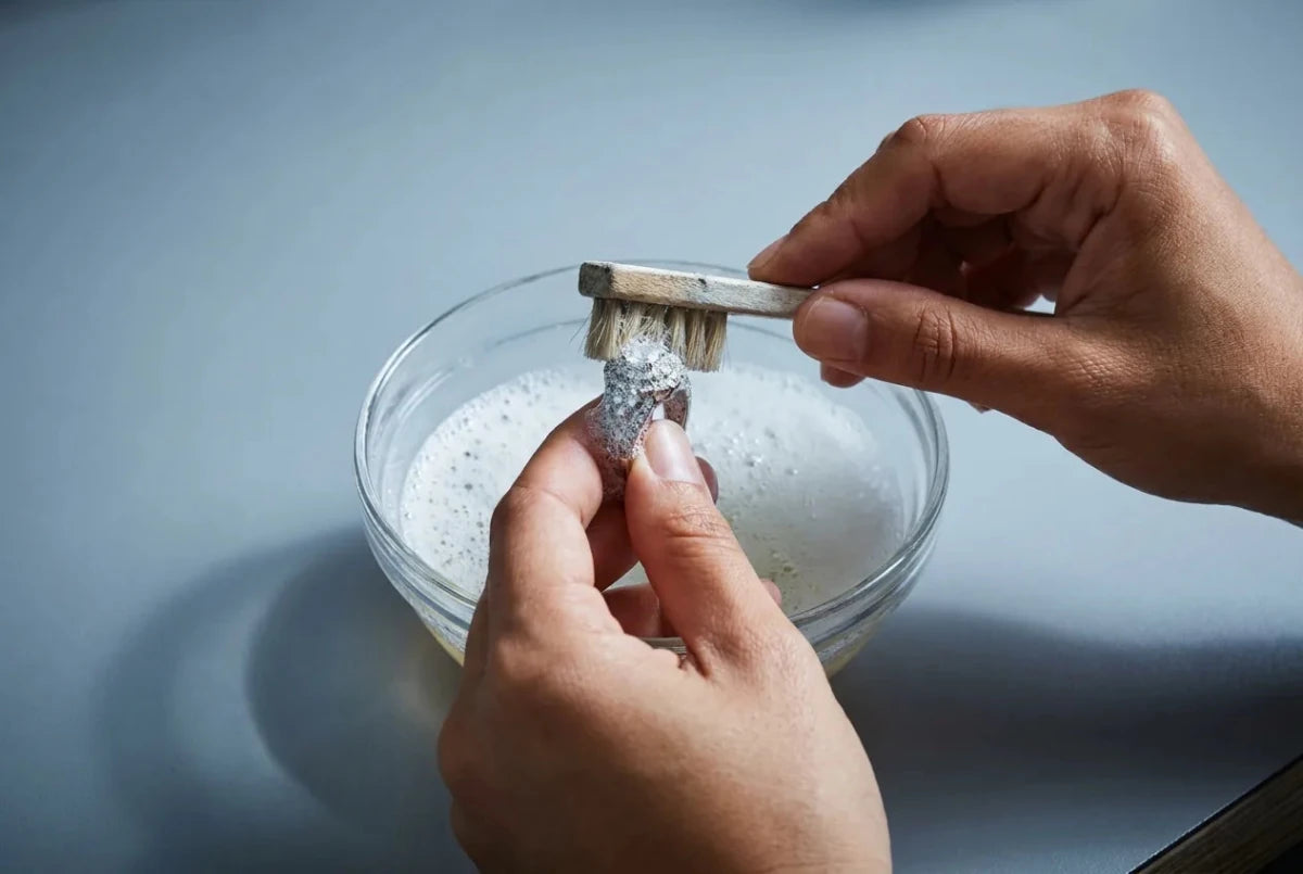 A jeweler's hands cleaning a white gold diamond ring with a soft brush over a bowl of soapy water