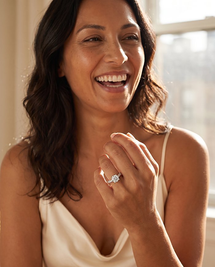A woman in cream silk laughing with joy, wearing a round-cut natural diamond engagement ring in white gold