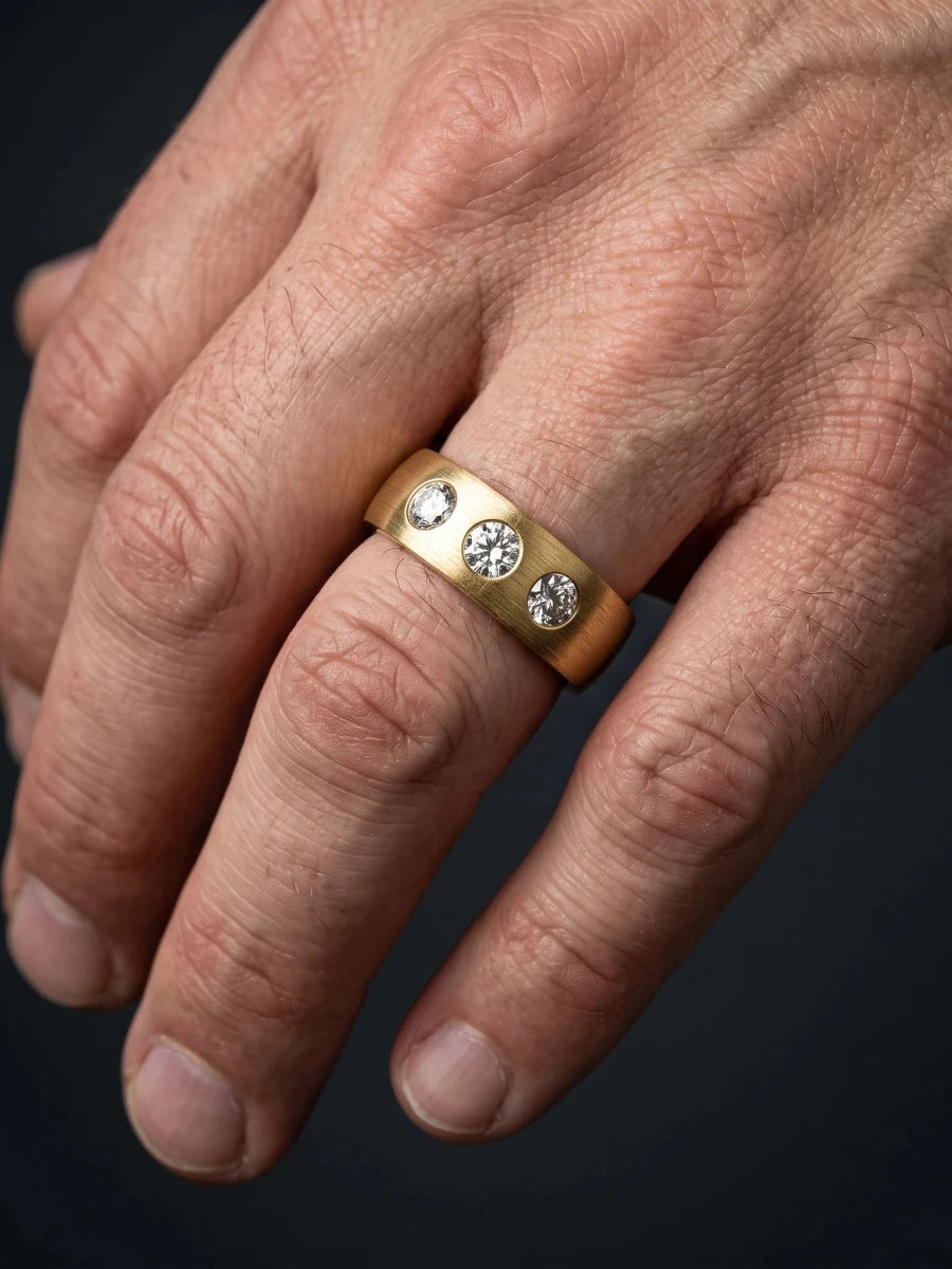 A wide brushed yellow gold men's ring with three flush-set diamonds, worn on a working hand against a dark slate background