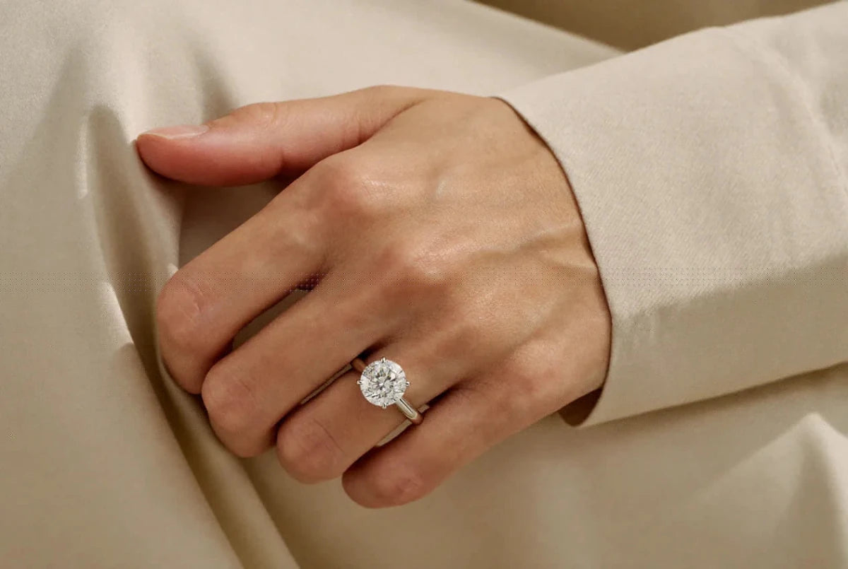 A round diamond solitaire in white gold worn on a hand resting against cream silk