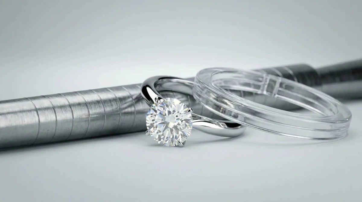 A white gold diamond ring beside a jeweler's ring mandrel and a plastic ring sizer on a studio surface