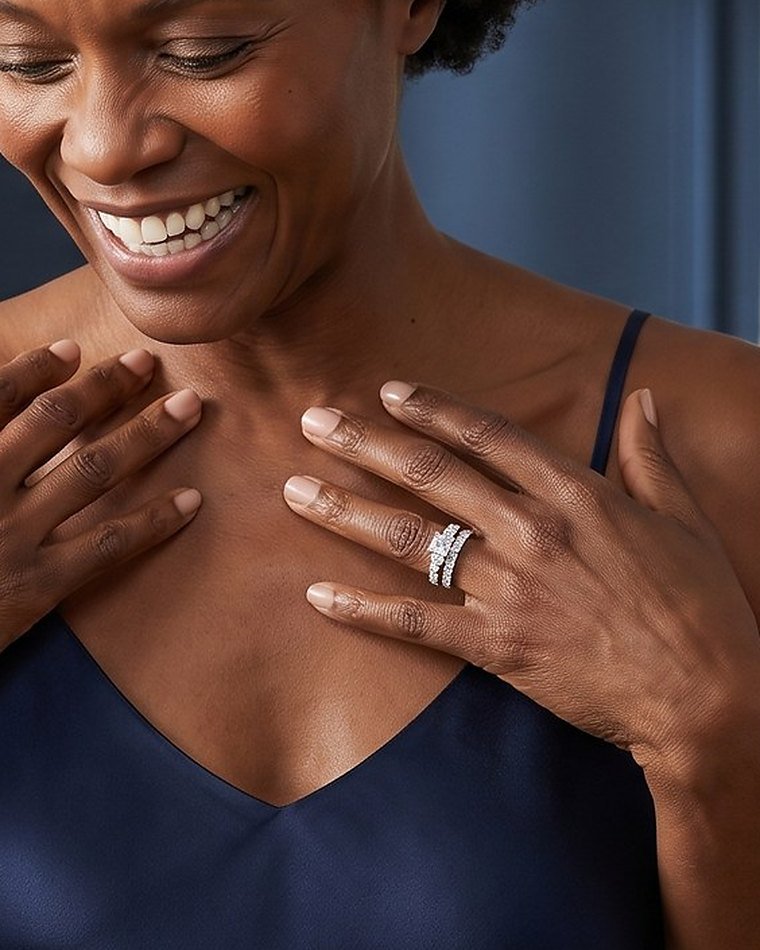 A smiling woman in a navy dress with her hand at her collarbone, wearing a diamond bridal set