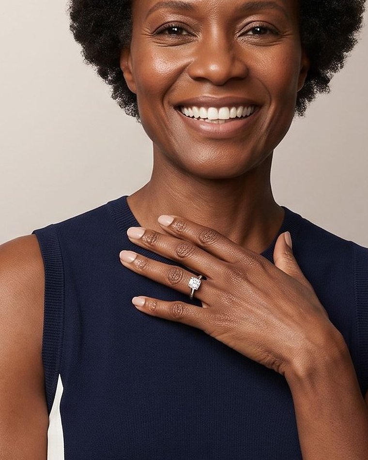 A laughing woman in a navy dress with her hand on her chest, wearing a solitaire diamond engagement ring