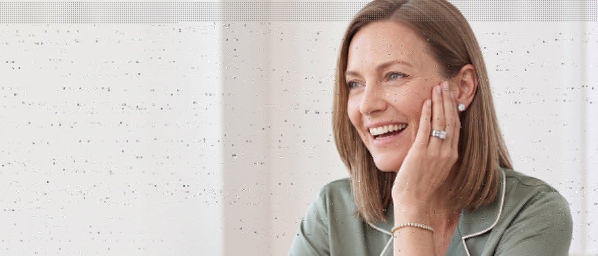 A laughing woman in a sage green silk top with her hand raised to her neck, wearing a diamond tennis bracelet, a diamond ring and a round diamond stud earring
