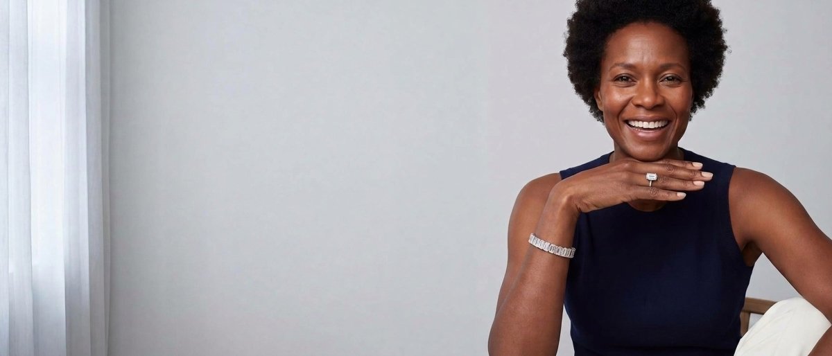 A smiling woman in a navy top with one hand resting at her collarbone, wearing an emerald cut diamond ring, an emerald cut diamond bracelet and emerald cut diamond stud earrings