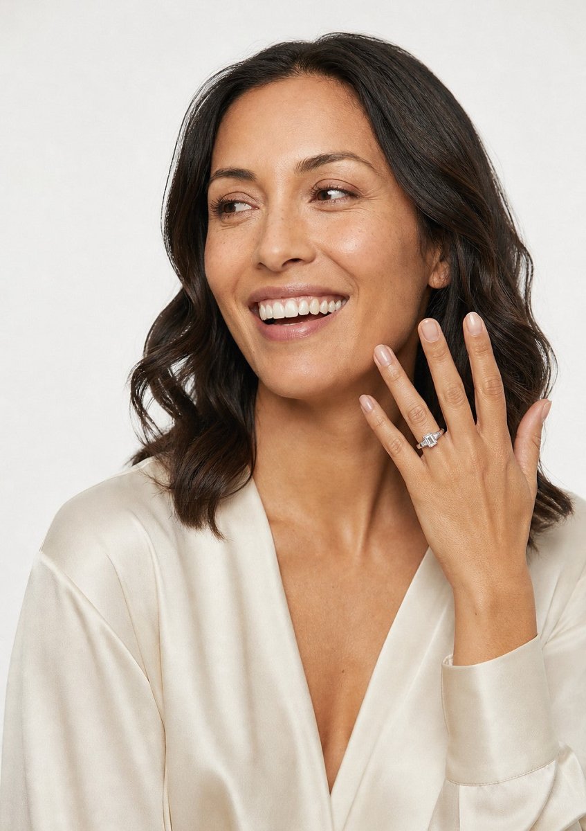 A laughing woman in cream silk with her hand raised near her collarbone, wearing a white gold emerald cut diamond solitaire engagement ring