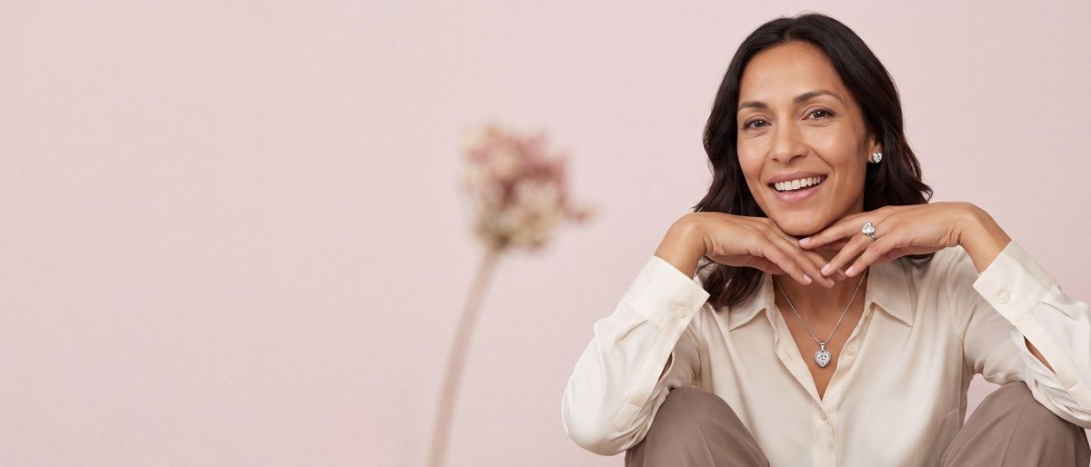 A smiling woman in a cream blouse with both hands framing her jaw, wearing a heart shaped diamond ring, a heart shaped diamond pendant and heart shaped diamond stud earrings