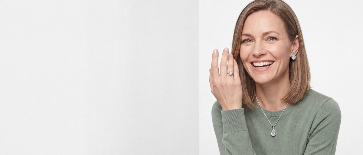 A smiling woman in a sage green sweater with one hand raised beside her face, wearing a pear shaped diamond ring, a pear shaped diamond pendant and pear shaped diamond stud earrings