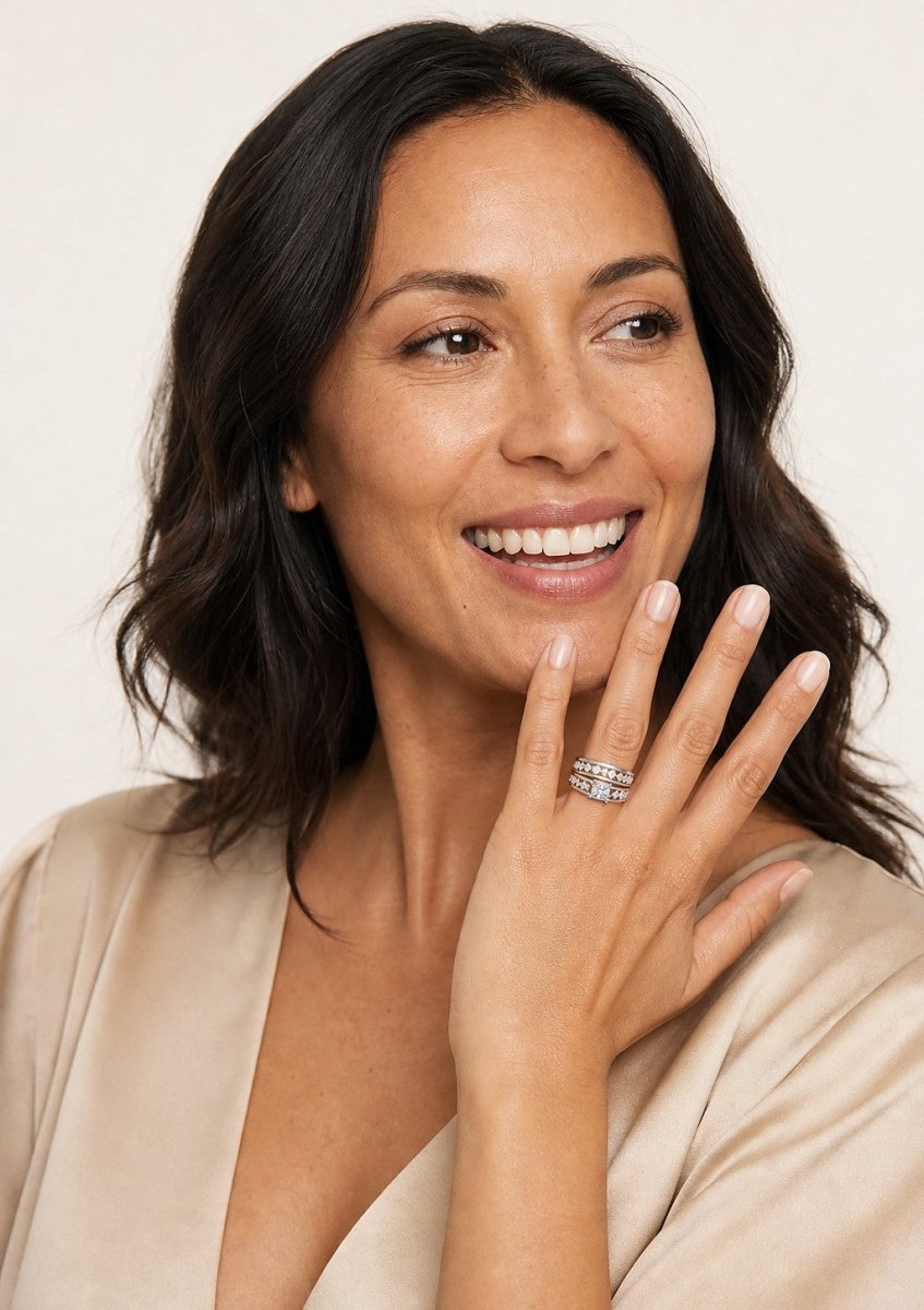 A laughing woman in cream silk with her hand curled beside her jaw, wearing a white gold princess cut diamond engagement ring on a channel set band