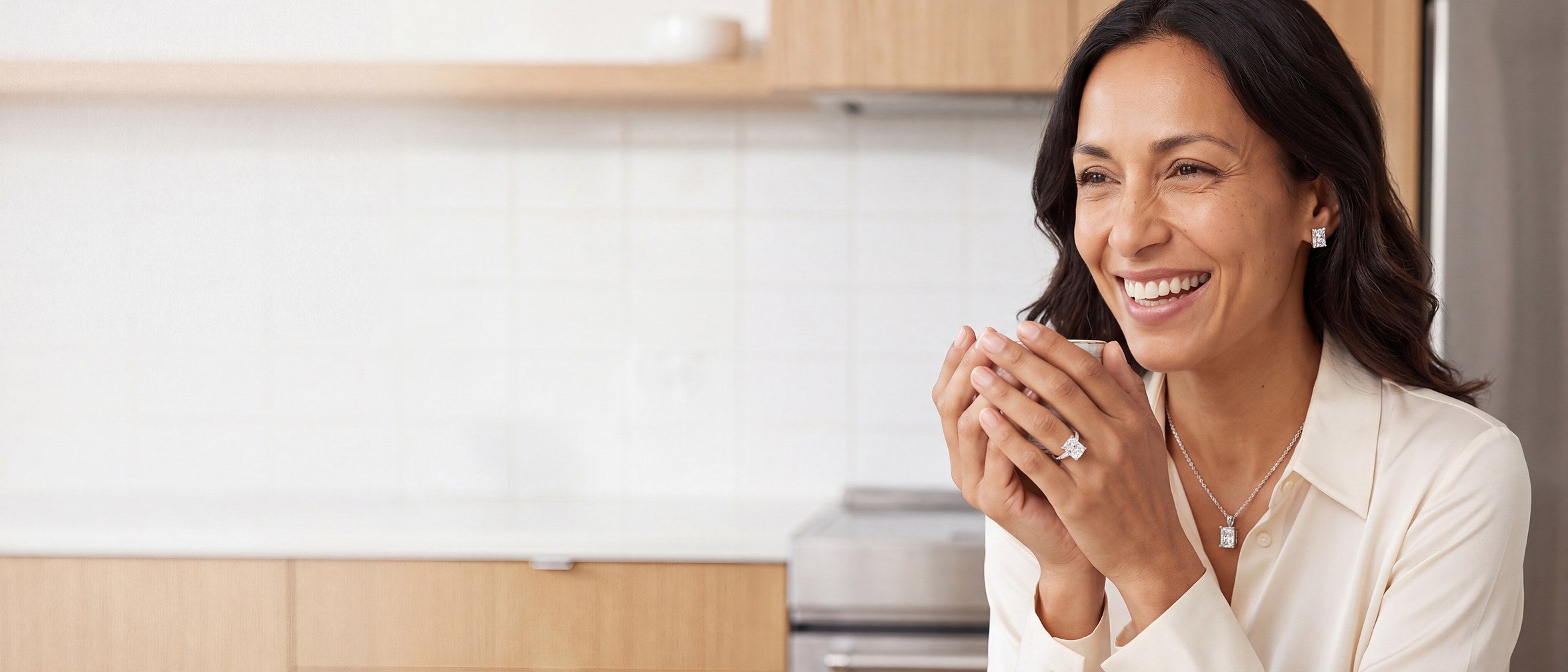 A smiling woman in a cream blouse holding a cup in both hands, wearing a radiant cut diamond ring, a radiant cut diamond pendant and radiant cut diamond stud earrings