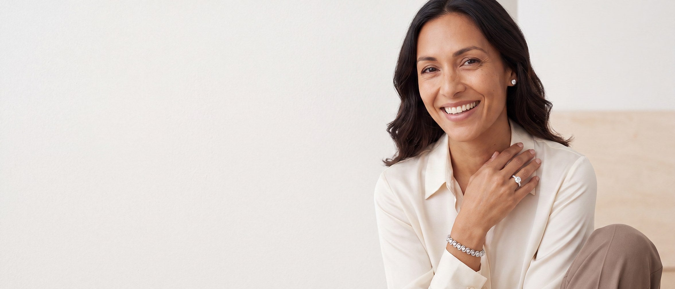 A laughing woman in a cream silk blouse with one hand raised to her collarbone, wearing a round diamond ring, a round diamond stud earring and a diamond tennis bracelet
