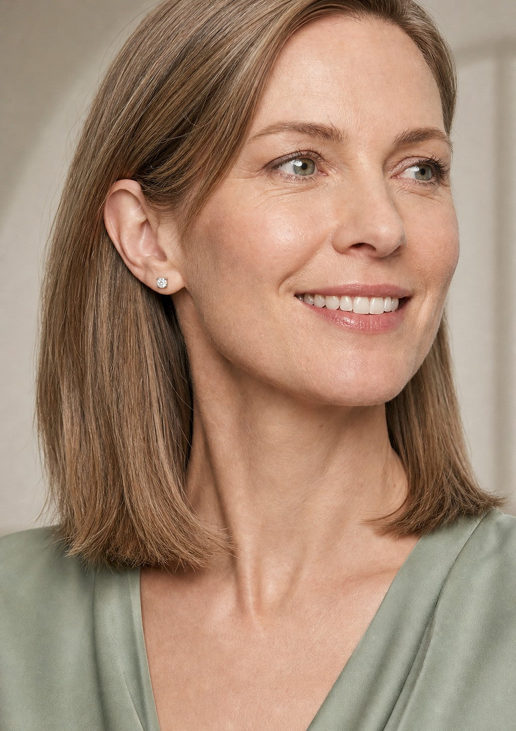 A smiling woman in sage green turned to show her ear, wearing a white gold round cut diamond stud earring