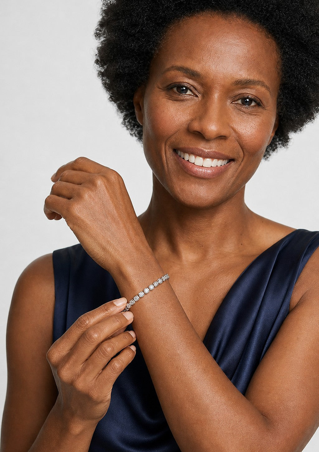 A smiling woman in navy holding her wrist to show a white gold round cut diamond tennis bracelet