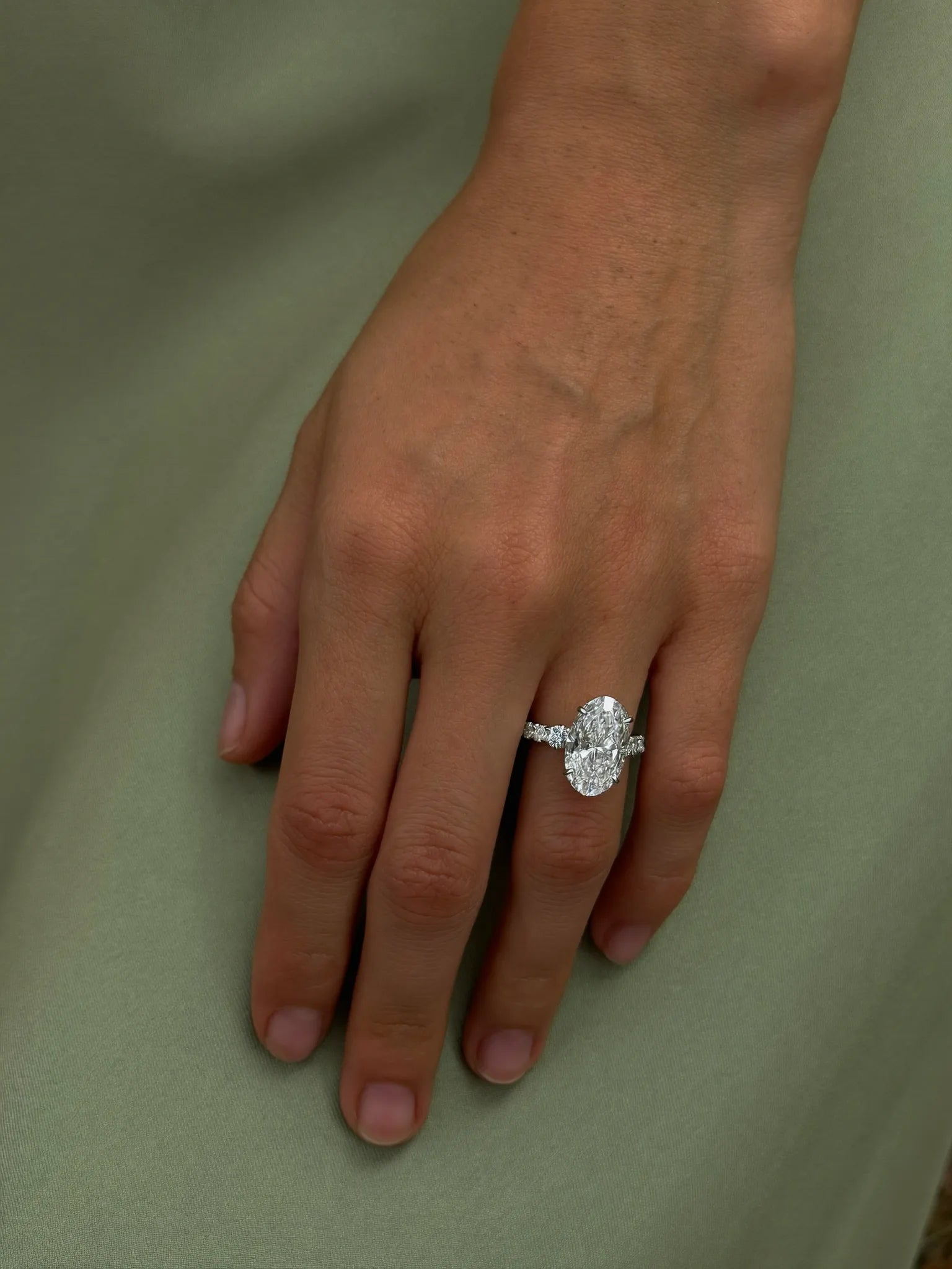A custom oval diamond engagement ring worn on her hand, resting on sage green silk